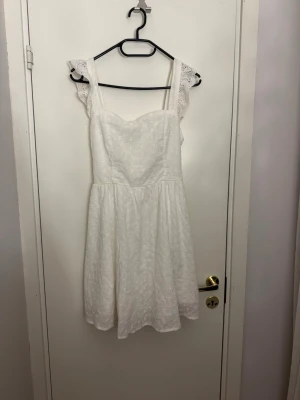 Vit spetsklänning med volang - Söt vit klänning med broderad spets, volangdetaljer på axelbanden och en stor rosett i ryggen. Klänningen är ärmlös med breda axelband. Perfekt för sommarens alla tillfällen.