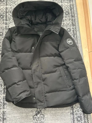 Svart Canada Goose jacka  - Säljer en svart dunjacka från Canada Goose med huva och dragkedja. Jackan har en klassisk patch på ärmen, två sidofickor och en varm, puffig design som passar perfekt för kalla vinter/vårdagar. Materialet är slitstarkt och jackan har en stilren look. Pris diskuteras och mer bilder via kontakt!