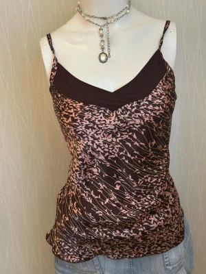 Brun och rosa blommig linnetopp - Snygg brun och rosa linnetopp med blommigt mönster och tunna axelband. Toppen har en v-ringad skärning och är tillverkad i ett glansigt, mjukt material som känns lyxigt mot huden. Perfekt att styla med jeans eller kjol för en trendig look.