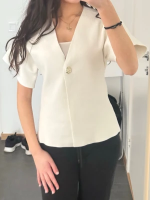 Vit kortärmad blus med knapp - Stilren vit blus med v-ringning och en dekorativ guldig knapp framtill. Blusen har korta, vida ärmar och rak passform. Materialet är tjockare och ger en exklusiv känsla. Perfekt att matcha med både jeans och kjol för en trendig look.