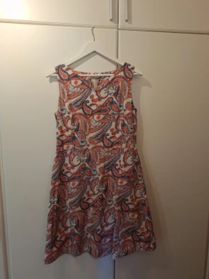 Mönstrad ärmlös kortklänning - Säljer en ärmlös kortklänning med färgstarkt paisleymönster i rött, blått och vitt. Klänningen har rund halsringning med litet v, dragkedja mitt bak samt lätt utställd kjol. Perfekt för dig som gillar 60-tals-vibbar och vill sticka ut med ett unikt mönster.