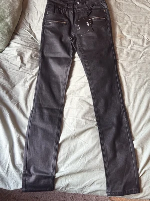 Svarta skin skinny jeans från FLEA - Snygga svarta skinny jeans från FLEA med glansig finish och dragkedjor på fickorna framtill. Jeansen har en smal passform och klassisk femficksdesign. Perfekta för dig som vill ha en edgy och trendig look.