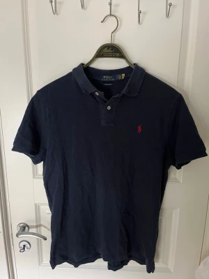 Marinblå piké från Polo Ralph Lauren - Klassisk marinblå pikétröja från Polo Ralph Lauren med röd broderad logga på bröstet. Modellen är custom slim fit och har korta ärmar samt knappar vid kragen. Perfekt för en stilren och avslappnad look. Storlek L i barn så är S i vuxenstorlek 