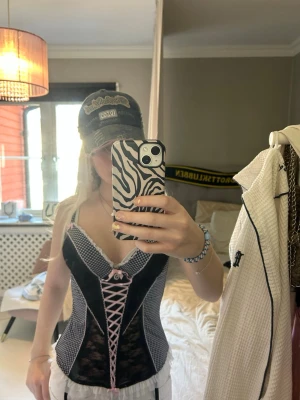 Svart och vit korsett med spetsdetaljer - Supersöt korsett i svart och vitt med rutigt mönster, spetsdetaljer och rosa snörning framtill. Dekorerad med små rosetter och volangkant upptill och nedtill. Axelbanden är justerbara och materialet är en mix av spets och tyg med stretch.