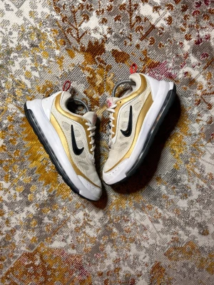Nike Air Max vita och guld sneakers - Nike Air Max sneakers i vitt och guld med svart swoosh på sidan. Ovandelen är i mesh och syntet, med vita skosnören och en chunky vit sula med svart yttersula. Röd detalj på hälen och AIR-tryck bak. Perfekta för dig som gillar streetwear och sportig stil.