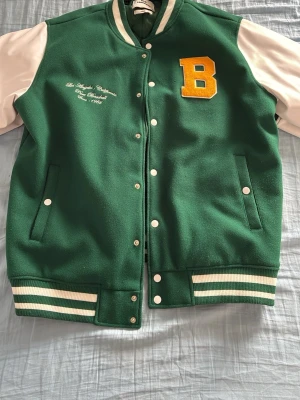 Grön bomberjacka med B-märke - Snygg grön bomberjacka med vita ärmar och gul B-patch på bröstet. Jackan har vita knappar, broderad text på vänster sida och randiga muddar i grönt och vitt. Insidan är quiltad i grönt satinliknande material. Perfekt för en sportig och retro look.