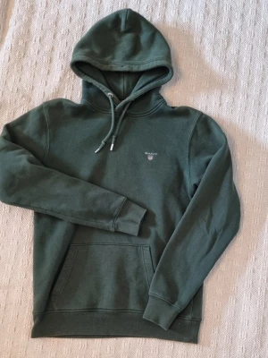 Mörkgrön hoodie från Gant - Snygg mörkgrön hoodie från Gant med broderad logga på bröstet. Klassisk känguruficka framtill, ribbade muddar och justerbar dragsko i huvan. Tillverkad i mjukt bomullsmaterial som känns skönt mot huden. Perfekt för en avslappnad och stilren look. Storlek small