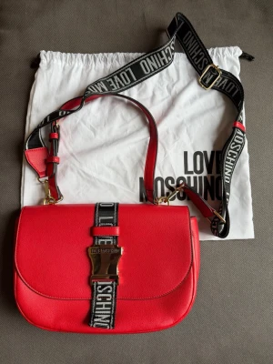 Love Moschino Väska - Röd handväska från Love Moschino med guldfärgat spänne. Justerbart axelband som kan tas av. Använd ca 3 gånger. Dustbag medföljer. 
