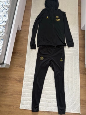 Svart PSG hoodie från Jordan - Säljer ett svart träningsset från PSG och Jordan med gul logga och Qatar Airways-tryck. Setet består av en hoodie med dragkedja och fickor samt matchande byxor. Inga skador, bra skick, bekväm och väldigt fin tracksuit 