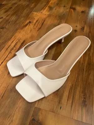 Vita slip-in pumps med låg klack - Säljer ett par stilrena vita slip-in pumps med öppen tå och fyrkantig framdel. Skorna har låg klack och bred rem över foten. Klacken är 5cm. Aldrig använda. Perfekta till studenten eller sommaren! 