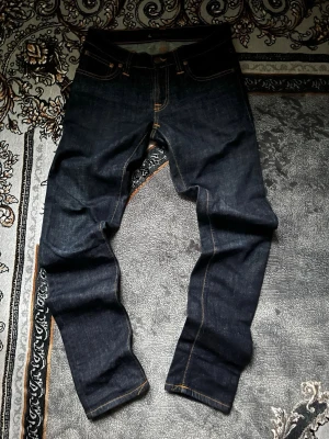 Mörkblå jeans från Nudie Jeans - Säljer ett par mörkblå jeans från Nudie Jeans i storlek 26/32. Klassisk femficksmodell med orange kontrastsömmar och rak passform. Tillverkade i kraftigt denimtyg som ger en snygg och tidlös look.