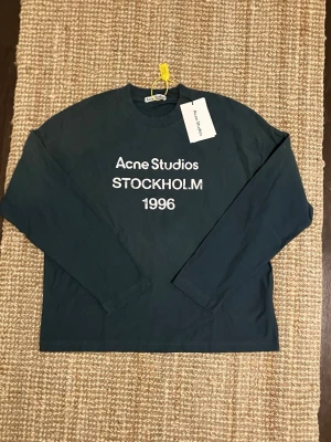 Mörkblå/svart långärmad topp Acne Studios - 100% äkta svart/mörkblå långärmad topp från Acne Studios med vit text 'Acne Studios STOCKHOLM 1996' på bröstet. Klassisk rund halsringning och relaxed fit. Perfekt för dig som gillar minimalistisk och stilren design. Pris kan sänkas vid en snabb affär. Jag säljer den för att den inte passar som jag velat så därför är den aldrig använd 