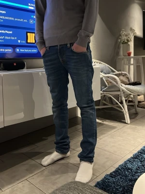 Nudie jeans - Säljer ett par blå slim fit (Grim Tim) jeans med snygga slitningar på bakfickorna och lätt tvättad look. Jeansen har klassisk femficksdesign och normal midja. Perfekta för dig som gillar en modern och avslappnad stil. 
