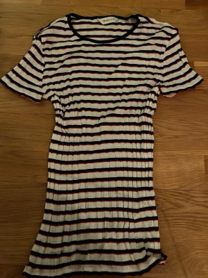 Randig kortklänning från Gant - Kortärmad ribbad klänning från Gant med svarta, vita och röda horisontella ränder. Klänningen har rund halsringning och är i en stretchig, mjuk bomullsblandning som sitter snyggt på kroppen.