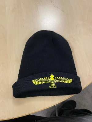 Svart mössa med gul vinge-tryck - Säljer en svart mössa med uppvikt kant och ett stort gult tryck av en stiliserad vinge och eld framtill. Mössan är ribbstickad och har en clean streetwear-look som passar perfekt till kalla dagar.