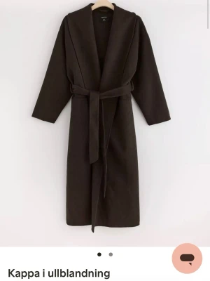 Mörkbrun kappa i ullmix från Uniqlo - Stilren mörkbrun kappa från Uniqlo i ullblandning. Modellen är lång med sjalkrage, raka ärmar och avtagbart midjebälte för en clean siluett. Perfekt för kyliga dagar och enkel att matcha med olika outfits. Det är hål i en av fickorna men går enkelt att sy ihop