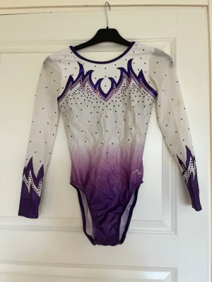 Lila och vit gymnastikdräkt med pärlor - Säljer en långärmad gymnastikdräkt i vitt och lila med snyggt ombre-mönster. Dräkten har dekorativa pärlor och glittriga detaljer över hela framsidan och ärmarna. Perfekt för träning eller tävling inom gymnastik.