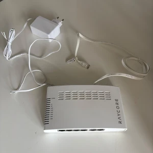 Raycore vit nätverksswitch 6 portar fiberdosa - Säljer en vit nätverksswitch från Raycore med sex LAN-portar och tillhörande strömadapter. Enheten är tillverkad i plast och har en stilren, rektangulär design med ventilationshål på ovansidan. Perfekt för att koppla ihop flera enheter i hemnätverket.