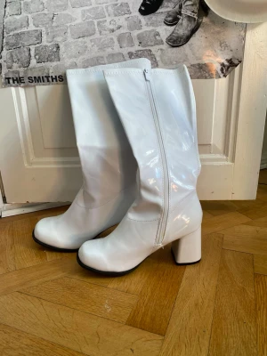 Vita lackstövlar, gogo boots - Säljer ett par höga vita stövlar i glansigt lackmaterial med chunky klack och dragkedja på sidan. Stövlarna har rundad tå och svart sula, perfekta för att sticka ut och ge retrovibbar till din outfit. Precis köpt dem på vinted, men säljer då de är för stora för mig. Pris kan diskuteras!