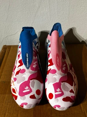 Adidas x BAPE F50 fotbollsskor - Unika Adidas x BAPE F50 fotbollsskor med färgstarkt camomönster i rosa, rött, vitt och blått. Skorna har en strömlinjeformad design utan snörning, med blå och rosa detaljer på plösen. Tillverkade i syntetmaterial för bästa prestanda på planen.