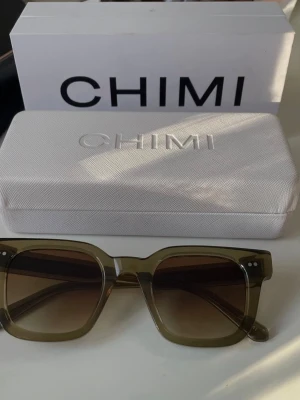 Olivgröna solglasögon från CHIMI - Snygga solglasögon från CHIMI med fyrkantig form därav modellen 04 och olivgröna bågar i syntetmaterial. Glasen är tonade i brunt och ger en cool vibe. Kommer med originaletui, putsduk och påse. Perfekta för soliga dagar och trendiga outfits.