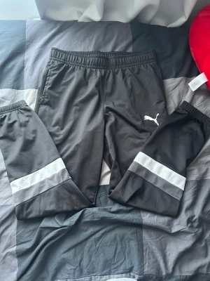 Svarta mjukisbyxor från Puma - Säljer ett par svarta mjukisbyxor från Puma med vita detaljer och reflexränder på benen. Byxorna har resår i midjan och är tillverkade i ett mjukt och stretchigt material som passar perfekt för chill eller träning. Klassisk Puma-logga på låret.