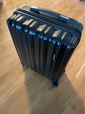 Svart kabinväska Regent med hjul - Snygg svart kabinväska från Regent med hårt skal och fyra smidiga hjul. Väskan har ett räfflat mönster, teleskophandtag i metall och bärhandtag upptill. Perfekt storlek för resor och enkel att rulla genom flygplatsen.