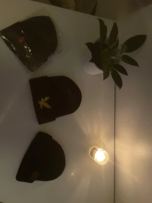 Svarta mössor med brodyr och patch - Tre svarta mössor i klassisk modell. En har gul broderad fågel, en har diskret brun patch framtill och en är förpackad i plast. Perfekt accessoar för kyliga dagar och enkel att matcha med olika outfits.