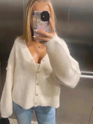 Vit fluffig kofta med knappar - Mysig vit kofta med v-ringning och stora silvriga knappar framtill. Koftan har en fluffig, mjuk känsla och avslappnad passform med långa ärmar. Perfekt att slänga över ett linne eller t-shirt för en chill look. Creme/vit