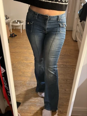 Blå bootcut jeans med fickdetaljer - Säljer ett par blå bootcut jeans med markerade sömmar och snygga fickdetaljer framtill. Jeansen har hög midja och bälteshällor, samt en lätt tvättad look. Perfekta för dig som gillar retrovibbar och vill ha ett par jeans som sticker ut.