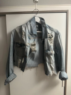 Ljusblå croppad distressed jeansjacka - Säljer en ljusblå croppad jeansjacka med slitna detaljer, rå kant och graffiti-liknande tryck på ryggen. Jackan har två bröstfickor med lock och knappar samt upprullade ärmar för en avslappnad streetstil.