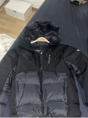 Svart dunjacka från Peak Performance - Snygg svart dunjacka från Peak Performance med huva och dragkedja framtill. Jackan har en puffig design, två sidofickor och logga på bröstet. Perfekt för kalla vinterdagar och riktigt varm tack vare dunfyllningen.