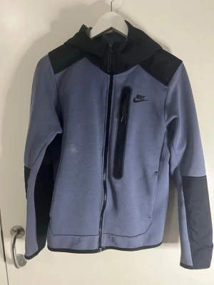Nike Tech Fleece hoodie blå/svart - Nike Tech Fleece hoodie i ljusblått med svarta detaljer och dragkedja framtill. Har en bröstficka med dragkedja och klassisk Nike-logga på bröstet. Tillverkad i mjukt och stretchigt material, perfekt för chill eller träning. Snygg huva och modern passform.