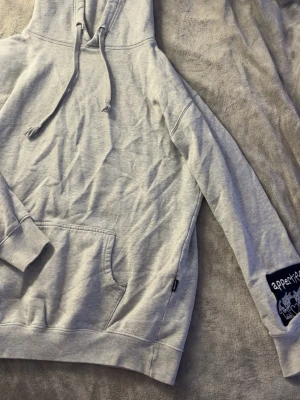 Grå hoodie från Appertiff med patch - Snygg grå hoodie från Appertiff med stor magficka och huva med snörning. På ena ärmen sitter en svart patch med vitt broderat motiv och texten 'appertiff'. Perfekt för en chill och avslappnad stil.