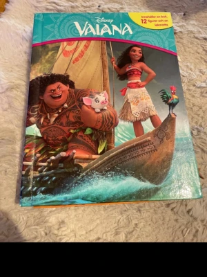 Disney Vaiana bok med figurer - Säljer en färgglad bok om Vaiana från Disney. Boken har hårda pärmar och medföljer 12 små figurer samt en lekmatta. Perfekt för dig som gillar äventyr och magiska berättelser. Omslaget visar Vaiana, Maui och deras djurvänner på en båt i havet.
