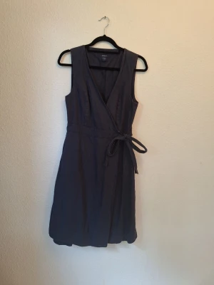 Mörkblå omlottklänning i linne från Gant - Snygg mörkblå omlottklänning från Gant med knytband i midjan och ärmlös design. Klänningen har v-ringning och är tillverkad i ett linne som känns skönt mot huden. Perfekt för en stilren och avslappnad look.