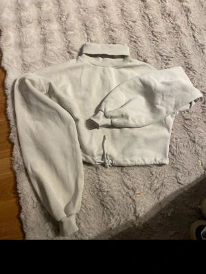 Vit croppad hoodie från NLY Trend - Säljer en vit croppad hoodie från NLY Trend med hög krage och dragsko i midjan. Hoodien har långa ärmar med ribbade muddar och är gjord i mjukt sweatshirtmaterial. Perfekt för en avslappnad och trendig look.