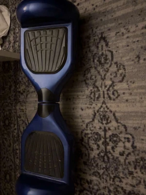 Blå hoverboard med svarta fotplattor - Säljer en blå hoverboard med svarta gummifotplattor. Hoverboarden har en modern design med rundade kanter och är tillverkad i plast och gummi. Perfekt för dig som vill ta dig fram smidigt och snabbt på ett kul sätt.