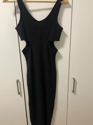 Svart ribbad långklänning med cut-outs - Trendig svart långklänning i ribbat material med v-ringning och markerade cut-outs i midjan. Klänningen är ärmlös och har en kroppsnära passform som framhäver siluetten. Perfekt för dig som vill ha en stilren men edgy look.