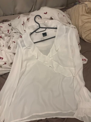 Vit volangblus från Vero Moda - Stilren vit blus från Gina tricot  med v-ringning och dekorativa volanger längs halsringningen. Blusen har lång ärm och är tillverkad i ett lätt, transparent tyg som ger en luftig känsla. Perfekt att matcha med jeans eller kjol för en trendig look.