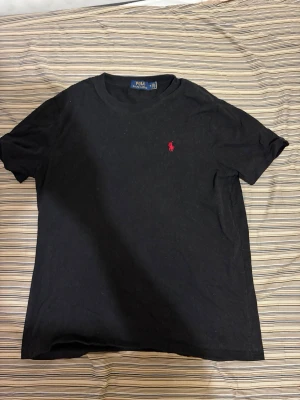 Svart t-shirt från Polo Ralph Lauren - Klassisk svart t-shirt från Polo Ralph Lauren med röd broderad logga på bröstet. T-shirten har rund halsringning och korta ärmar. Tillverkad i mjuk bomull för en skön känsla och enkel stil. Sitter pefekt som S
