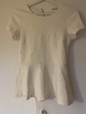 Vit peplumtop med strukturmönster - Säljer en vit peplumtop med korta ärmar och snyggt strukturmönster i tyget. Toppen har rund halsringning och dragkedja bak. Perfekt för dig som gillar feminina detaljer och en lite mer dressad look.