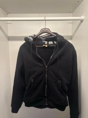 Svart hoodie från Burberry - Svart hoodie från Burberry med dragkedja, två stora fickor framtill och klassisk huva med snören. Insidan av huvan har Burberrys rutmönster i svart, vitt och rött. Broderad logga på bröstet. Mjuk bomullsblandning för skön känsla.