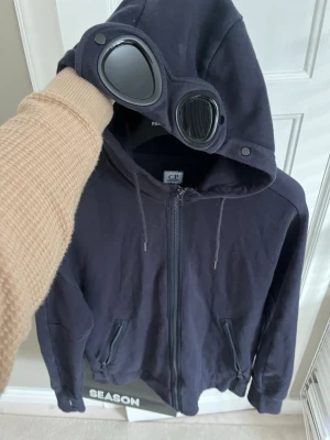 Svart hoodie med glasögon från C.P. Company - Säljer en mörkblå hoodie från C.P. Company med dragkedja framtill och två fickor. Huvan har inbyggda glasögon och snörning, vilket ger en unik och cool look. Perfekt för dig som vill sticka ut med streetwear-vibe. Tillverkad i mjukt bomullsmaterial.