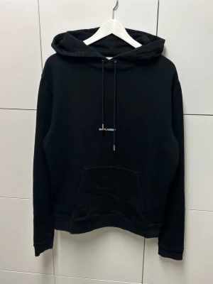 Saint Laurent Paris Hoodie  - Snygg svart hoodie från Saint Laurent med minimalistisk logga på bröstet. Klassisk känguruficka framtill, justerbar dragsko i huvan och mjukt material som känns skönt mot huden. Perfekt för en clean och stilren look.