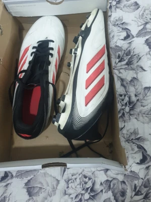 Adidas vita och röda fotbollsskor - Säljer ett par klassiska Adidas fotbollsskor i vitt med röda ränder och svarta detaljer. Skorna har dobbar för bra grepp på planen och snörning framtill. Insidan är röd och yttermaterialet är syntetiskt för slitstyrka och lätt vikt. Perfekta för dig som vill sticka ut på fotbollsplanen.