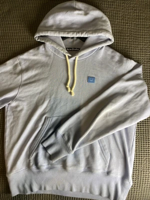 Ljuslila hoodie från Acne Studios - Snygg ljuslila hoodie från Acne Studios med klassisk känguruficka och huva med vita snören. På bröstet sitter Acne Studios ikoniska ansiktslogga i blått. Tillverkad i mjuk bomull för maximal komfort och stilren look.