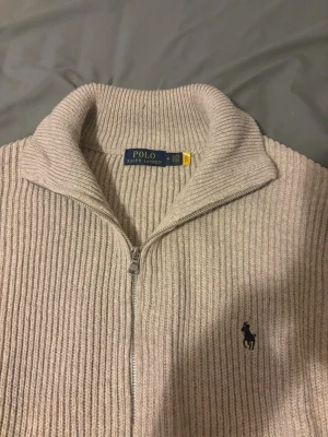 Beige ziptröja från Polo Ralph Lauren - Ribbstickad beige ziptröja från Polo Ralph Lauren med hög krage och dragkedja framtill. Tröjan har det klassiska mörkblå logobroderiet på bröstet och är tillverkad i mjukt material som passar perfekt för lager på lager.