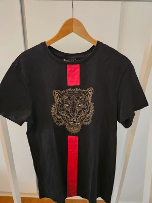 Svart t-shirt med tigertryck Closure - Svart t-shirt från Closure London med ett stort tigertryck i guld på bröstet och en röd vertikal rand som går genom trycket. Klassisk rund hals och korta ärmar. Perfekt för dig som vill sticka ut med en cool och edgy look.