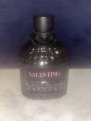 Tom Valentino born in roma  svart parfymflaska - Säljer en lyxig parfym från Valentino i en svart, mönstrad glasflaska med nitar och elegant design. Flaskan har en rund form och ett svart lock. Märkets logga syns tydligt i rosa på framsidan. Perfekt för dig som gillar exklusiva dofter och snygg inredningsdetalj.